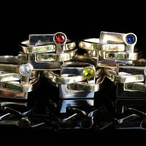 May include: Five silver rings with different coloured gemstones. The rings are stacked on top of each other and are reflected in a black surface. The gemstones are red, blue, clear, green, and light blue.