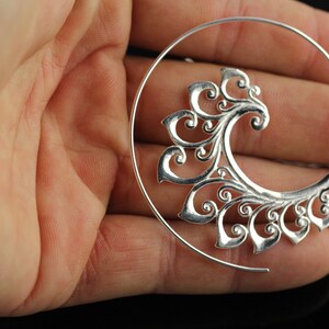 May include: Silver hoop earring with a decorative, swirling, floral design. The earring is open and has a circular shape.