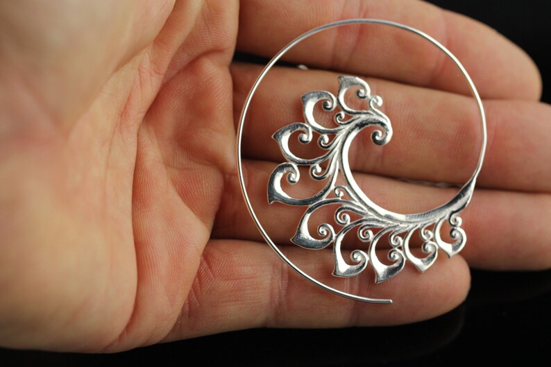 May include: Silver hoop earring with a decorative, swirling, floral design. The earring is open and has a circular shape.