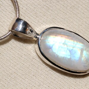 May include: A silver chain necklace with a pendant featuring a large oval-shaped moonstone. The moonstone has a milky white color with iridescent blue and green flashes.