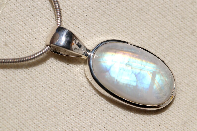May include: A silver chain necklace with a pendant featuring a large oval-shaped moonstone. The moonstone has a milky white color with iridescent blue and green flashes.