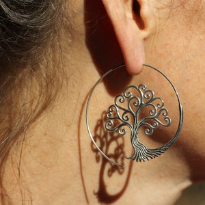 May include: Silver hoop earrings featuring a tree of life design. The tree is crafted from intricate swirls and has a detailed trunk.