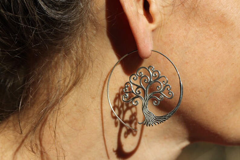 May include: Silver hoop earrings featuring a tree of life design. The tree is crafted from intricate swirls and has a detailed trunk.