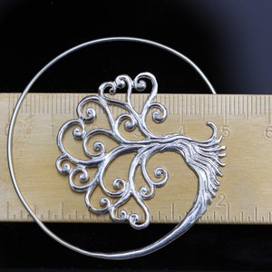 May include: Silver hoop earrings featuring a tree of life design. The tree is crafted from delicate swirls and branches.