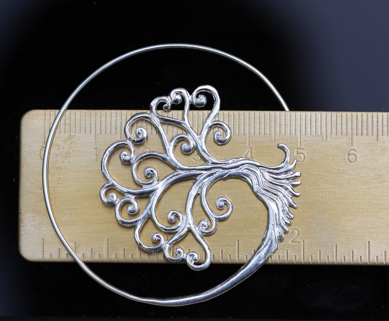 May include: Silver hoop earrings featuring a tree of life design. The tree is crafted from delicate swirls and branches.