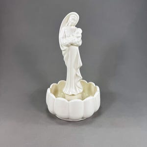 May include: A white ceramic figurine of the Virgin Mary holding baby Jesus, standing above a scalloped-edge bowl. The religious statue is a devotional item, likely for home decor or religious practice.