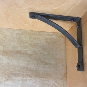 Puede incluir: Soporte de estante de metal negro con un brazo curvo, montado en una superficie de madera. El soporte tiene varios tornillos visibles.