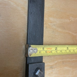 Puede incluir: Primer plano de una barra de metal negro con una pieza cuadrada de metal en el extremo. Una cinta m&eacute;trica amarilla se coloca sobre la barra, mostrando una medida de 1 pie o 38,5 cm.