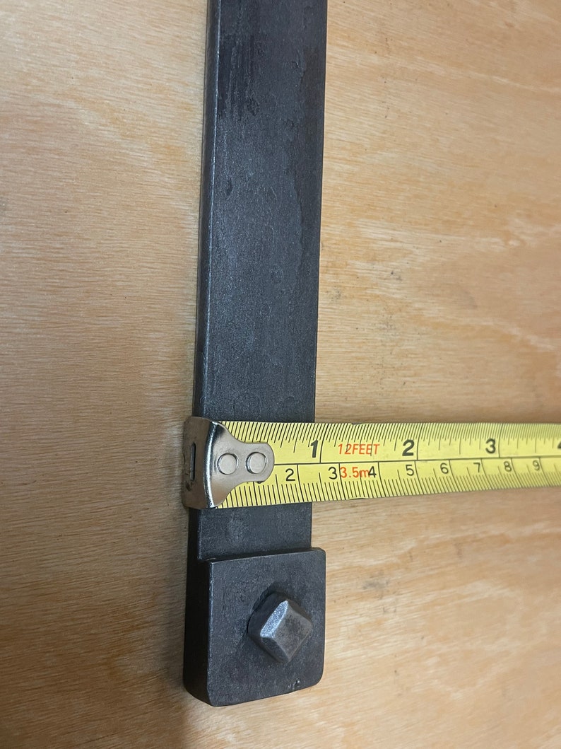 Puede incluir: Primer plano de una barra de metal negro con una pieza cuadrada de metal en el extremo. Una cinta m&eacute;trica amarilla se coloca sobre la barra, mostrando una medida de 1 pie o 38,5 cm.