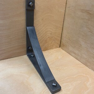 Puede incluir: Un soporte de estante de metal negro con forma triangular y borde curvo. El soporte tiene cuatro remaches cuadrados en la parte superior e inferior.