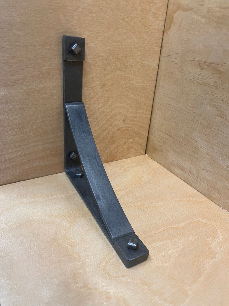 Puede incluir: Un soporte de estante de metal negro con forma triangular y borde curvo. El soporte tiene cuatro remaches cuadrados en la parte superior e inferior.