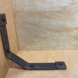 Puede incluir: Una escuadra de esquina de metal oxidado con cuatro tornillos negros. La escuadra est&aacute; unida a una superficie de madera.