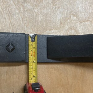 Puede incluir: Primer plano de un soporte de metal negro con una placa cuadrada y una barra rectangular. Se utiliza una cinta m&eacute;trica amarilla para medir el soporte, que tiene 7,6 cm de largo.