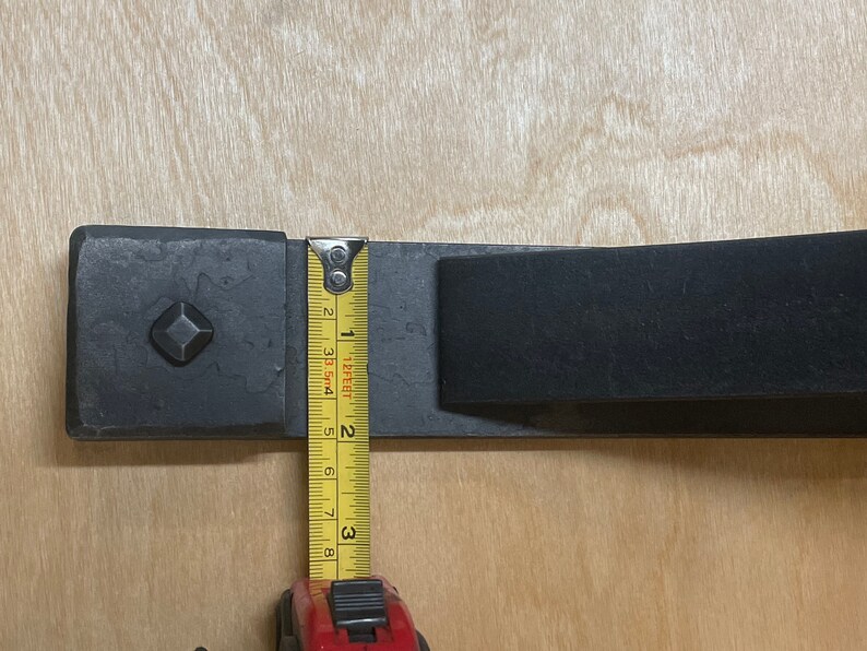 Puede incluir: Primer plano de un soporte de metal negro con una placa cuadrada y una barra rectangular. Se utiliza una cinta m&eacute;trica amarilla para medir el soporte, que tiene 7,6 cm de largo.