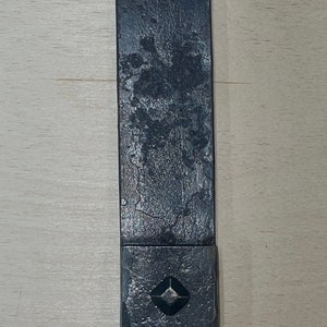 Puede incluir: Una correa de metal negro con una pieza de metal cuadrada en el extremo. La pieza de metal tiene un recorte en forma de diamante en el centro.