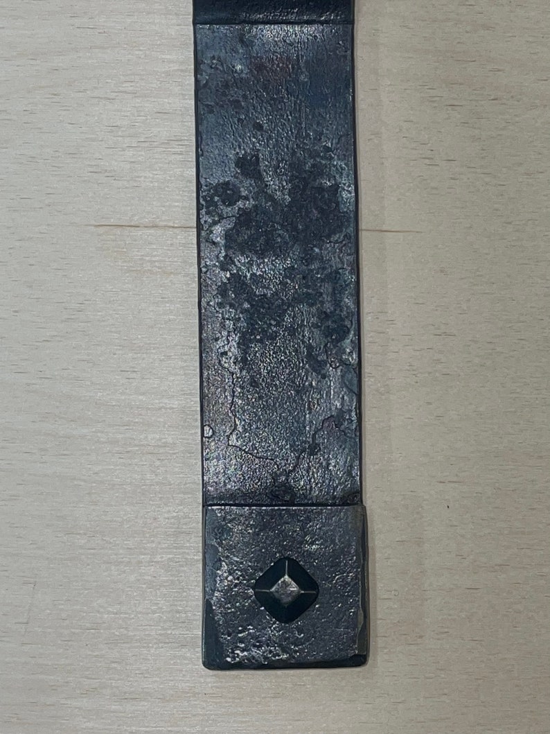Puede incluir: Una correa de metal negro con una pieza de metal cuadrada en el extremo. La pieza de metal tiene un recorte en forma de diamante en el centro.