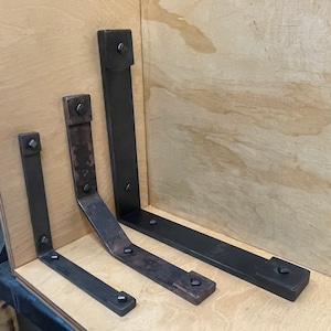 Puede incluir: Cuatro soportes de estante de metal negro de diferentes tama&ntilde;os, montados en una superficie de madera clara. Los soportes tienen forma de L y tienen un acabado decorativo ligeramente desgastado.