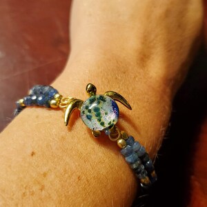 Op de afbeelding: Een armband met een goudkleurige schildpadbedel met een glazen schild. De schildpad heeft een blauw, groen en goudkleurig ontwerp. De armband heeft blauwe kralen en gouden accenten.