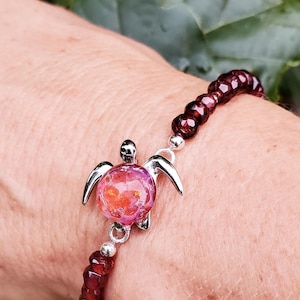 Könnte beinhalten: Ein Armband mit einem Schildkröten-Anhänger. Das Armband besteht aus dunkelroten Perlen und hat einen Schildkröten-Anhänger mit einem rosa-orangenen Zentrum und silbernen Akzenten. Das Armband wird am Handgelenk getragen.
