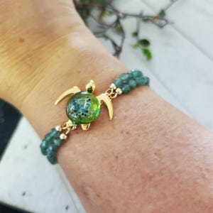 Puede incluir: Una pulsera con dije de tortuga dorada con un centro de vidrio verde y azul. La pulsera presenta una doble hebra de pequeñas cuentas verdes. El dije de tortuga tiene un diseño detallado y es el punto focal de la pulsera.