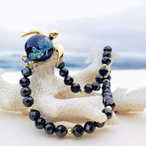 Puede incluir: Una pulsera de cuentas negras con un dije de tortuga dorada. El dije de tortuga es azul oscuro con detalles dorados. La pulsera se exhibe sobre un objeto blanco similar al coral.