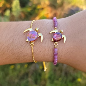 Puede incluir: Dos pulseras con colgantes de tortuga doradas. Una pulsera tiene una cadena, la otra tiene un diseño de cuentas. Los colgantes de tortuga tienen una concha de resina morada y rosa. El fondo es verde borroso.