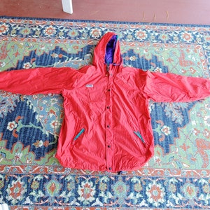 May include: A red hooded jacket with a purple lining and a zipper closure. The jacket has two pockets on the front and a drawstring hood.