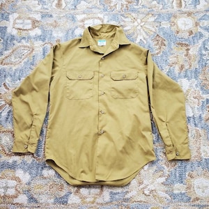 May include: A long-sleeved, button-down shirt in a muted mustard yellow color. The shirt has two chest pockets with button closures and a curved hem.