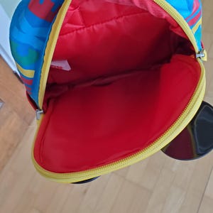 May include: A colorful backpack with a red interior and a yellow zipper. The exterior features a vibrant pattern of blue, red, and yellow. The backpack is open, revealing the spacious interior.
