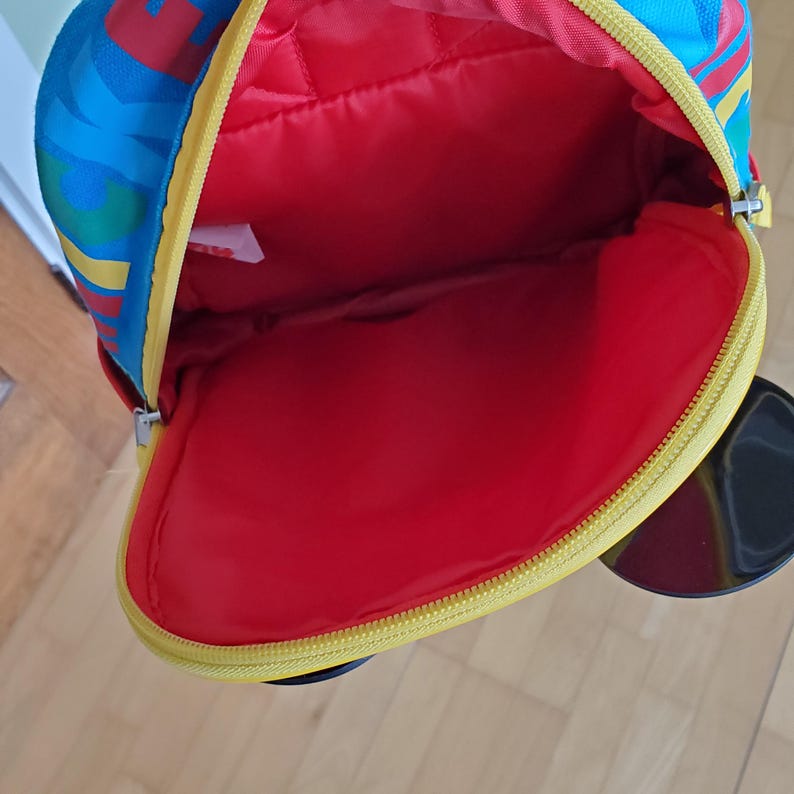 May include: A colorful backpack with a red interior and a yellow zipper. The exterior features a vibrant pattern of blue, red, and yellow. The backpack is open, revealing the spacious interior.