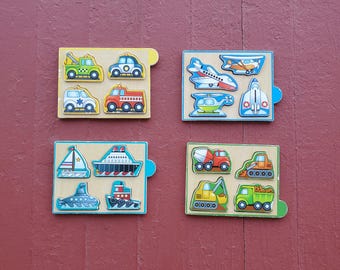 May include: Four wooden puzzles with transportation themes. Each puzzle features a different set of vehicles: cars, airplanes, boats, and construction vehicles. The puzzles are on a red surface.