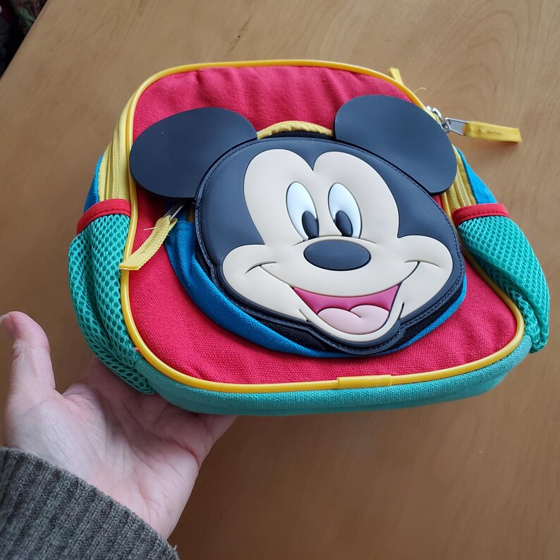 May include: A colorful backpack featuring a large Mickey Mouse face. The backpack has a red body, blue accents, and yellow trim. It includes a mesh side pocket and a zipper closure. The Mickey Mouse face is black, white, and pink.