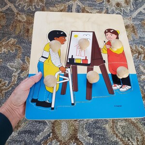 May include: A wooden puzzle with five knobs for children to remove and replace. The puzzle depicts two people, one using a walker and the other drawing on an easel. The puzzle is on a blue background with the word "Kaplan" in the bottom right corner.