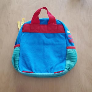 May include: A small, blue backpack with a red handle and trim. The backpack has two side pockets made of green mesh. The top of the backpack has a red strap. The backpack is approximately 10 inches tall and 8 inches wide.
