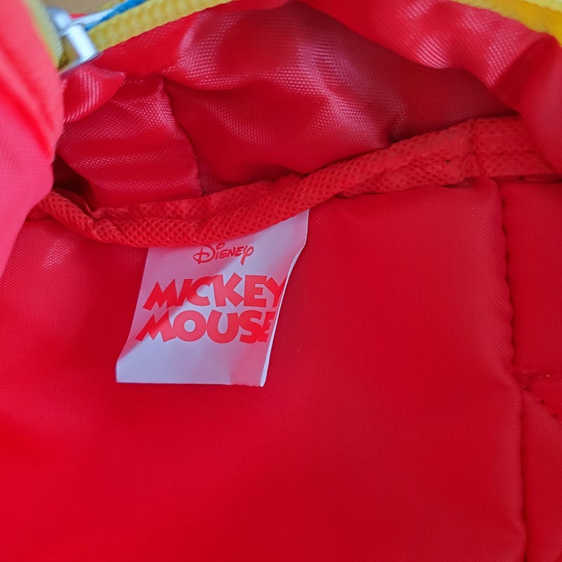 May include: Red interior of a Disney Mickey Mouse backpack. The interior is red with a white tag that reads "Disney Mickey Mouse" in red and white lettering. The backpack has a yellow and blue zipper.