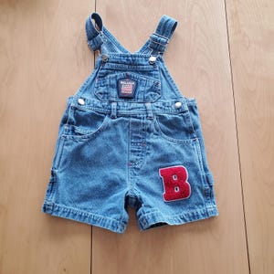 May include: Blue denim baby overalls with adjustable straps and pockets. The overalls feature a red patch with the letter "B" and a small "Beluga Jeans" label. The overall shorts are designed for a child.