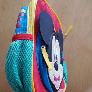 May include: A colorful backpack featuring Mickey Mouse. The backpack has a red body with blue and yellow accents. A mesh pocket is on the side. The zipper is yellow, and the Mickey Mouse face is a 3D design.