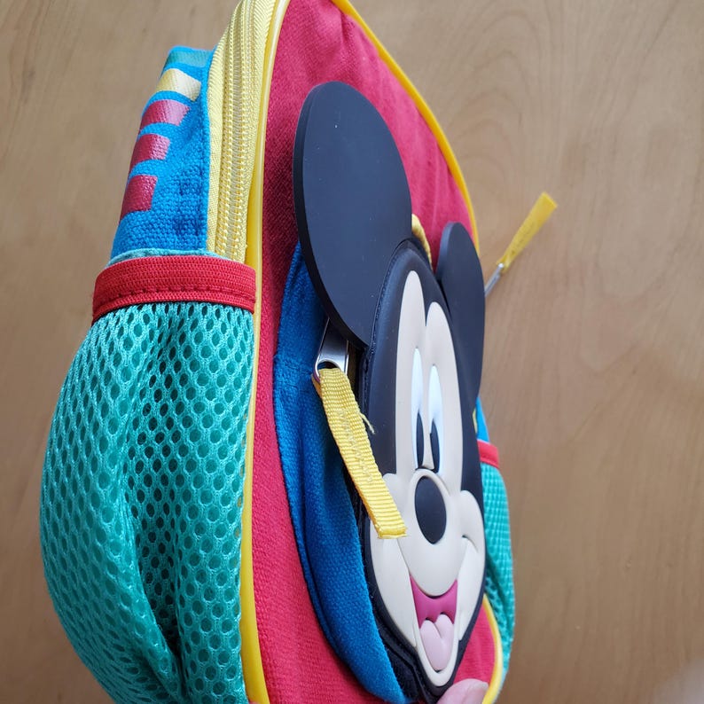 May include: A colorful backpack featuring Mickey Mouse. The backpack has a red body with blue and yellow accents. A mesh pocket is on the side. The zipper is yellow, and the Mickey Mouse face is a 3D design.