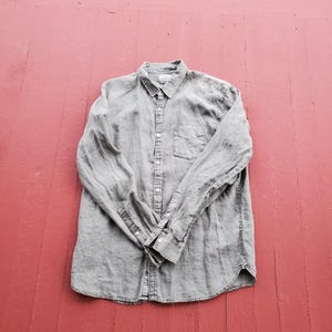 May include: A gray button-down shirt with long sleeves, a chest pocket, and a collar. The shirt is made of a soft, textured fabric and is laid flat on a red surface. The shirt has a classic design.