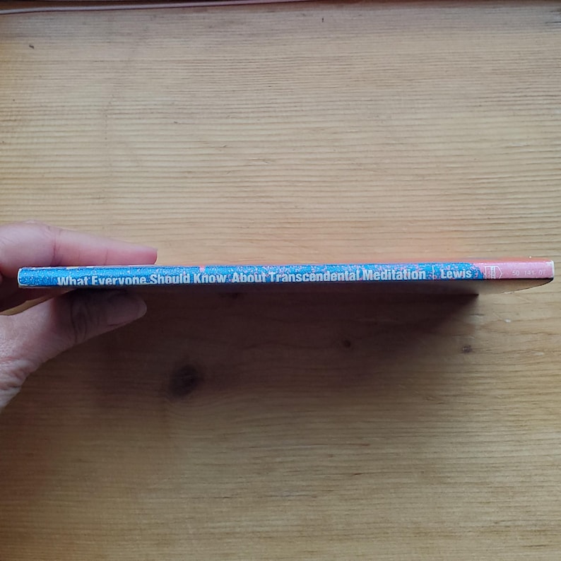 May include: A book titled "What Everyone Should Know About Transcendental Meditation" by Lewis. The book's cover is blue with white text and pink accents. The book is held by a person.