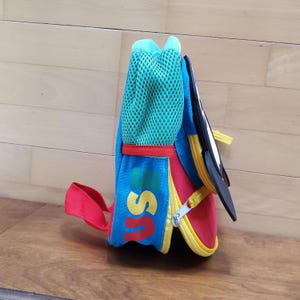 May include: A colorful backpack with a blue body, red accents, and a yellow zipper. The backpack features a green mesh pocket and the letters "US" in yellow. A black and white graphic is attached to the side.