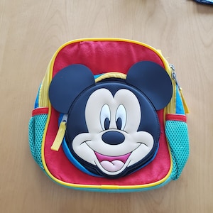 May include: A colorful backpack featuring Mickey Mouse's face. The backpack has a red body with yellow and blue accents, black ears, and a smiling Mickey face. It includes a zippered main compartment and mesh side pockets.