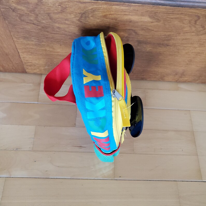 May include: A colorful backpack featuring the text "Mickey" in red and yellow on a blue background. The bag has a red strap, yellow accents, and black circular details resembling ears. The bag is open, revealing a yellow interior.