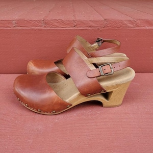 May include: A pair of brown leather clogs featuring a rounded toe, a wide strap across the instep, and an adjustable ankle strap with a metal buckle. The footwear has a light-colored wooden heel and sole with visible metal studs along the base.