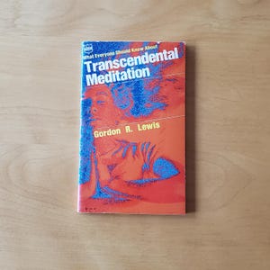 May include: A book titled "What Everyone Should Know About Transcendental Meditation" by Gordon R. Lewis. The cover is orange with blue and red graphics, featuring a person's silhouette. The book is on a light brown wooden surface.