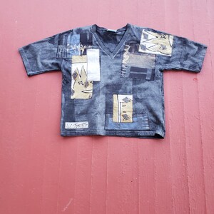 May include: A short-sleeved, V-neck shirt in shades of blue with patchwork designs. The patches feature geometric and abstract patterns in beige, white, and navy. The text "N.CTORIAS" is visible near the bottom. The shirt is laid flat on a pink surface.