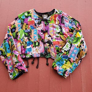 May include: A colorful cropped jacket with a patchwork design featuring various floral and geometric patterns. The jacket has a button closure and a drawstring waist.