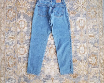 May include: Light blue denim jeans with a classic five-pocket design. The jeans feature a straight leg cut and a small red tag on the back pocket. The jeans are laid flat on a patterned rug.