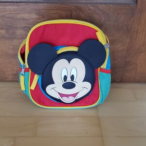 May include: A red, blue, and yellow backpack featuring a large Mickey Mouse face on the front. The backpack has a zippered main compartment, mesh side pockets, and adjustable straps. The backpack is designed for children.