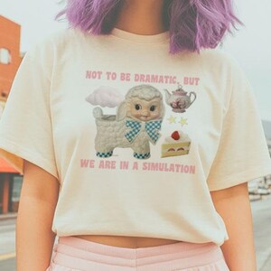 May include: A woman with purple hair is wearing a pale yellow t-shirt with a cartoon lamb and the text "Not to be dramatic, but we are in a simulation". She is also wearing pink sweatpants.